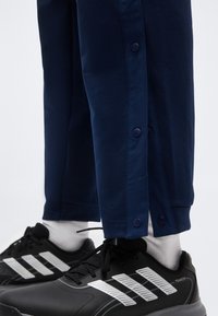 Κοντινό πλάνο σκούρου μπλε παντελονιού με κουμπιά στο πλάι και μαύρα αθλητικά παπούτσια με λευκές ρίγες και λευκές κάλτσες.