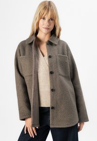 Vrouw met lang blond haar, gekleed in een knoopjesjasje van textuur in taupe boven een beige V-hals trui en donkere spijkerbroek, staand tegen een witte achtergrond.