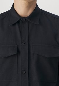 Black button-up shirt with a collared neck, featuring two chest pockets and a textured fabric. Buttons are black and rounded.
