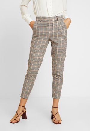 Femme portant un pantalon à carreaux beige, noir et rouge, fuselé avec des plis, associé à un chemisier blanc à manches longues et des sandales à talons carrés marron.