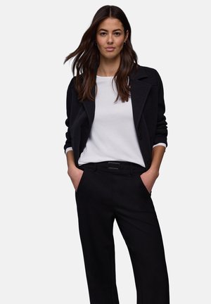 Veste noire en mélange de laine avec col cranté, associée à une chemise blanche en coton à manches longues et un pantalon noir, avec un détail ceinture.