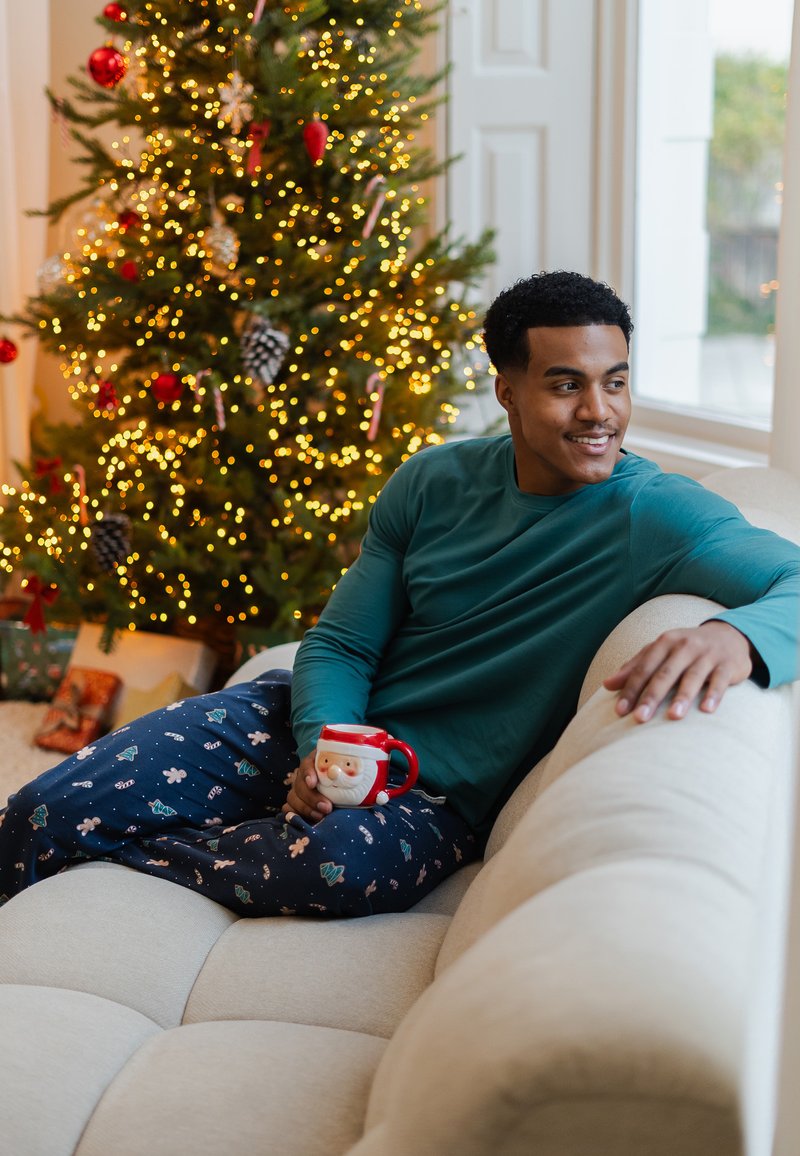 Gezellig tealkleurig lang mouw shirt en marineblauwe pyjamabroek met feestelijke patronen. Houdt een rood mok met een afbeelding van de Kerstman vast, zittend op een beige bank. Feestelijke boom op de achtergrond.