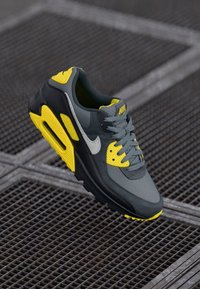 Gray Nike sneaker with yellow accents and a gray swoosh, floating above a textured surface, showcasing its design and colors.