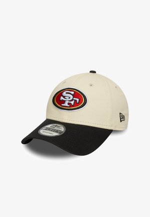 Beige baseball cap with a black bill, featuring an embroidered red and white logo on the front and an adjustable strap at the back.