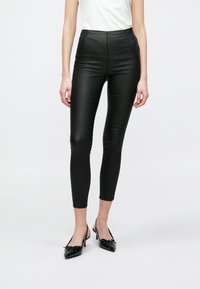 Fekete műbőr leggings, magas derékkal és sima, fényes textúrával. Testhez álló dizájn, amely a bokáig szűkül. Fekete magassarkú cipővel párosítva.