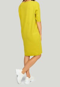 Żółta sukienka z krótkim rękawem o luźnym kroju, sięgająca do połowy uda, zestawiona z białymi sneakersami. Gładka tekstura, brak wzorów lub zdobień.
