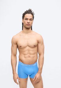 Homme torse nu avec des dreadlocks portant un boxer bleu Tommy Hilfiger, debout devant un fond blanc uni.