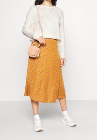 Pull en maille crème, jupe midi à rayures orange avec des accents jaunes et blancs, et baskets blanches, associés à un petit sac bandoulière beige.