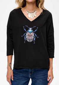 Woman wearing a black V-neck shirt with a large blue beetle graphic and a silver feather necklace, paired with blue jeans and visible tattoos on hands.