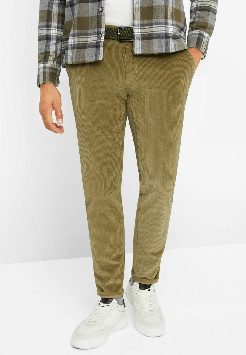 Homme portant un pantalon en velours côtelé vert olive, une chemise à carreaux verts et gris, une ceinture noire et des baskets blanches sur un fond clair.