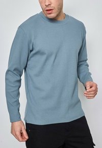 Chemise bleu clair à manches longues en coton texturé, avec un design simple, des poignets côtelés et un ourlet droit.