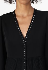 Robe noire avec un décolleté en V profond orné de clous en argent. Le tissu a une texture lisse, avec des détails plissés à la taille.