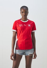 Sarkans futbols krekls ar baltiem akcentiem, Šveices karoga emblēma, Puma logo un vertikālām svītrām. Īsas piedurknes ar kontrastējošiem dizaina elementiem.