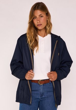 Femme portant une veste zippée marine sur une chemise blanche boutonnée, jean bleu et ceinture marron, tenant les bords de la veste devant un fond clair.