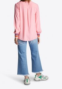Camicia rosa a maniche lunghe con polsini a righe, abbinata a jeans a gamba larga di jeans chiaro con orlo sfrangiato e sneakers beige con dettagli verdi.