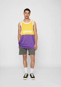 Lilla og gul utenermet skjorte med "STARTER"-logo, kombinert med grå shorts. Modellen har på seg svarte sko med kremfargede såler og hvite sokker.