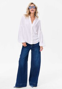 Witte blouse met opgestoken mouwen, gecombineerd met blauwe wijde jeans. De outfit heeft een casual en losse pasvorm, gemaakt van zachte stoffen.