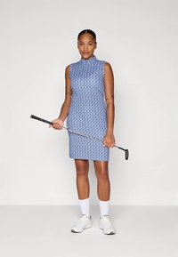 Γυναίκα με αμάνικο μπλε φόρεμα γκολφ με σχέδια, κρατώντας μια γκολφ μπαστούνι, φορώντας λευκά αθλητικά παπούτσια και κάλτσες, στέκεται μπροστά σε απλό φόντο.