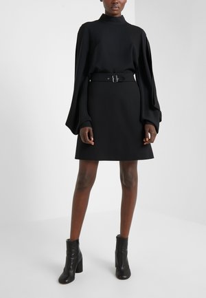 Frau trägt ein schwarzes, langärmeliges Kleid mit Gürtel in der Taille und schwarze Stiefeletten, steht vor einem schlichten Hintergrund.
