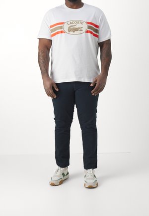 T-shirt en coton blanc avec de larges rayures horizontales rouges et marron, arborant un logo Lacoste. Assorti à un pantalon marine foncé et des baskets multicolores.