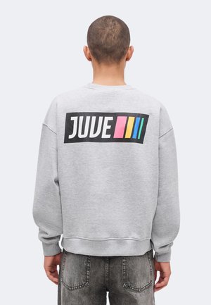 Persoană cu păr scurt poartă un hanorac gri cu textul „JUVE” și dungi roz, galbene, albastre pe spate, asortat cu blugi negri decolorați.