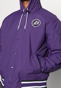 Lila puffjacka med hög krage, vita dragsnören, sidofickor och randig svart- och vit nederkant. Har en cirkulär logotyp på bröstet.
