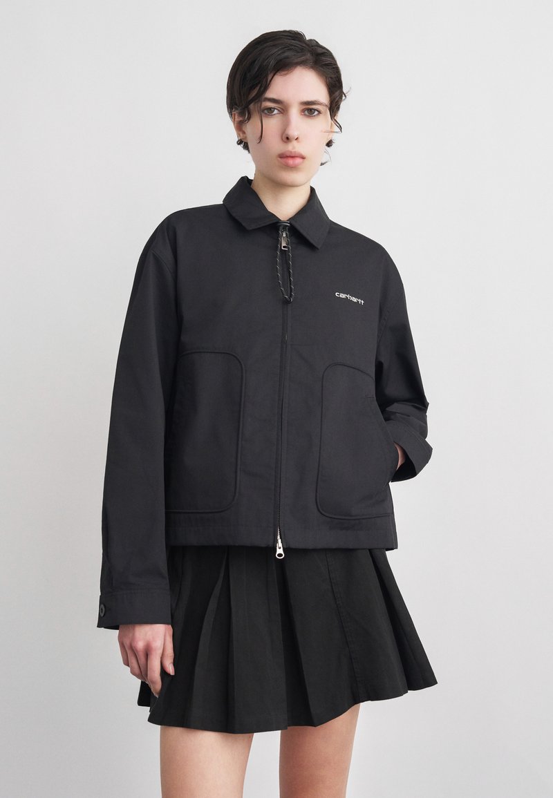 Νεαρό άτομο με κοντά σκούρα μαλλιά φοράει μαύρο μπουφάν με φερμουάρ και πλισέ φούστα, στέκεται μπροστά από απλό φωτεινό φόντο.