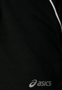 Top de sport noir avec une texture lisse, une bande d'accentuation blanche et un logo argenté placé en bas à droite.