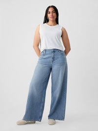 Woman with braided hair wearing a white sleeveless top, wide-leg light blue jeans, and beige sneakers standing with hands behind back.