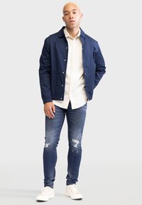 Chaqueta azul marino con cierre de botones a presión sobre una camisa crema de botones, combinada con jeans azul desgastados y zapatillas blancas. Tejido suave y diseño ajustado.