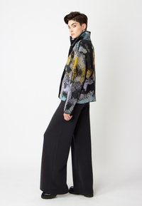 Vícebarevná vlněná bunda s abstraktními vzory v žluté, modré a černé barvě; spárovaná s černými kalhotami s vysokým pasem a širokými nohavicemi a černými botami.