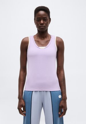 Čierna osoba nosiaca fialový top bez rukávov cez biele tielko a modré nohavice širokého strihu s vertikálnymi čiernymi pruhmi.