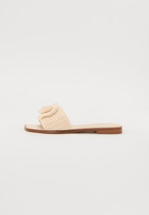 Beige crochet slide sandal with a woven strap and a flat, brown sole. Simple design, soft texture, and a rounded bow detail on the strap.
