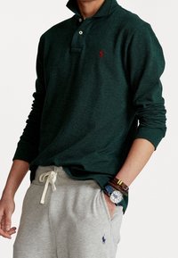 Grünes Langarm-Polohemd mit Kragen und zwei Knöpfen, versehen mit einem roten Logo. Kombiniert mit grauen Jogginghosen und geschichteten Armbändern.