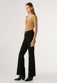 Bodysuit de manga larga en color beige con un escote en V profundo, combinado con jeans acampanados negros de cintura alta, llevados con botas de tacón negras hasta el tobillo.