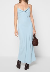 Femme portant une robe nuisette bleu clair sans manches avec des bretelles fines, tenant un sac à main marron foncé et portant des bottes pointues marron foncé.