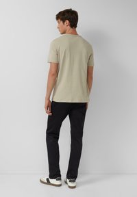 Mężczyzna odwrócony plecami, ubrany w beżowy t-shirt z krótkim rękawem, czarne spodnie i białe sneakersy na jednolitym jasnoszarym tle.