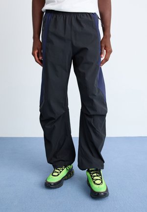 Black trousers with navy side panels, elastic waistband, and zipper pockets, paired with green sneakers featuring black accents.