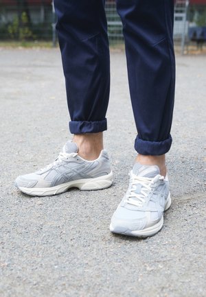 Persona con pantalones azul marino remangados y zapatillas Asics gris claro de pie sobre un camino de grava.