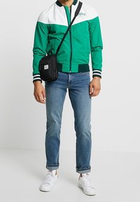 Zielona i biała kurtka bomberka z zamkiem z przodu, ze ściągaczami w paski, w połączeniu z jasnoniebieskimi dżinsami i białymi trampkami oraz czarną saszetką na ramię.