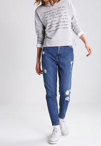 Sudadera gris claro con texto estampado, combinada con jeans de mezclilla azul desgastados. Zapatillas blancas completan el look casual.