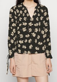 Blusa nera con stampa floreale beige, scollatura a V e maniche lunghe con dettagli annodati. Indossata con pantaloni corti rosa chiaro.