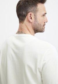 Lys beige, langermet skjorte med rund hals, som har en ribbet tekstur. Baksiden viser en subtil sømdetalj og en synlig tatovering.