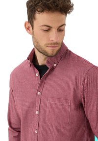 Uomo con capelli ricci corti che indossa una camicia a quadretti rossi e bianchi sopra una maglietta nera, che guarda verso il basso a sinistra.