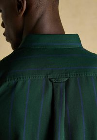 Nahaufnahme eines Halses und einer Schulter einer Person, die ein dunkelgrünes Hemd mit dünnen blauen Streifen trägt. An dem Hemd befindet sich ein kleines Stoffetikett mit der Aufschrift "Joules".