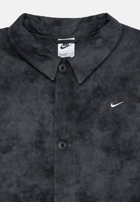 Nike skjorte i svart stoff med et teksturert mønster, med spiss krage, to knapper og en liten hvit logo på venstre side.