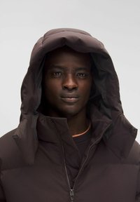 Ruskea pehmustettu takki, jossa on korkea kaulus ja suuri hupullinen kaula-aukko; edessä vetoketju ja nappikoristeet. Pehmeä, sileä tekstuuri.