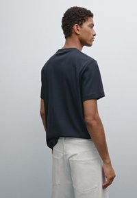 Uomo di lato che indossa una maglietta nera a maniche corte e pantaloni bianchi su uno sfondo chiaro uniforme.
