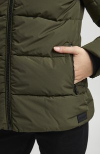 Grön puffjacka med quiltat mönster, ficka med dragkedja och svart dragkedja. Har en logotyp-patch vid fållen och en figurnära design.