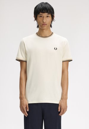 Homme portant un t-shirt à manches courtes écru avec un bord noir et doré et un petit logo laurier, debout devant un fond uni.